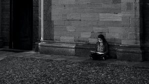 In this black and white image, a person is sitting on a cobblestone street with their back against a brick wall reading a book.