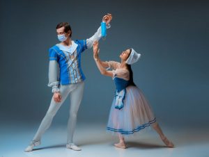 Photo is of two ballet dancers. The male dancer is wearing a blue face mask and dangling a blue face mask over the body of the female dancer who is posed as if she is pleading to him. Both dancers are wearing costumes of blue and white shirts and white dancing tights.