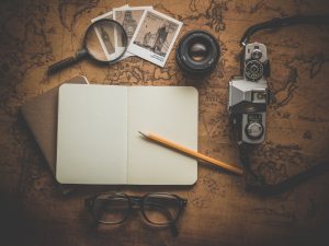 This is a photo of a journal open to a blank page with a pencil lying atop it. Surrounding the journal are a magnifying glass, a pair of eyeglasses, a camera, and several old-fashioned looking photographs.