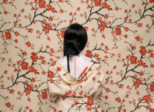 A woman is standing in front of a wall with her back turned to the camera. The woman is wearing a kimono that with flowers on it that matches the flowers of the wallpaper on the wall. This makes the woman look like she is blending into the wallpaper.
