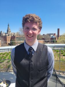 The writer is standing on a deck or porch that has a metal railing behind him. He is wearing glasses and has short, curly hair. He is also wearing a dress vest and tie. The writer is smiling with his mouth open looking directly at the camera.