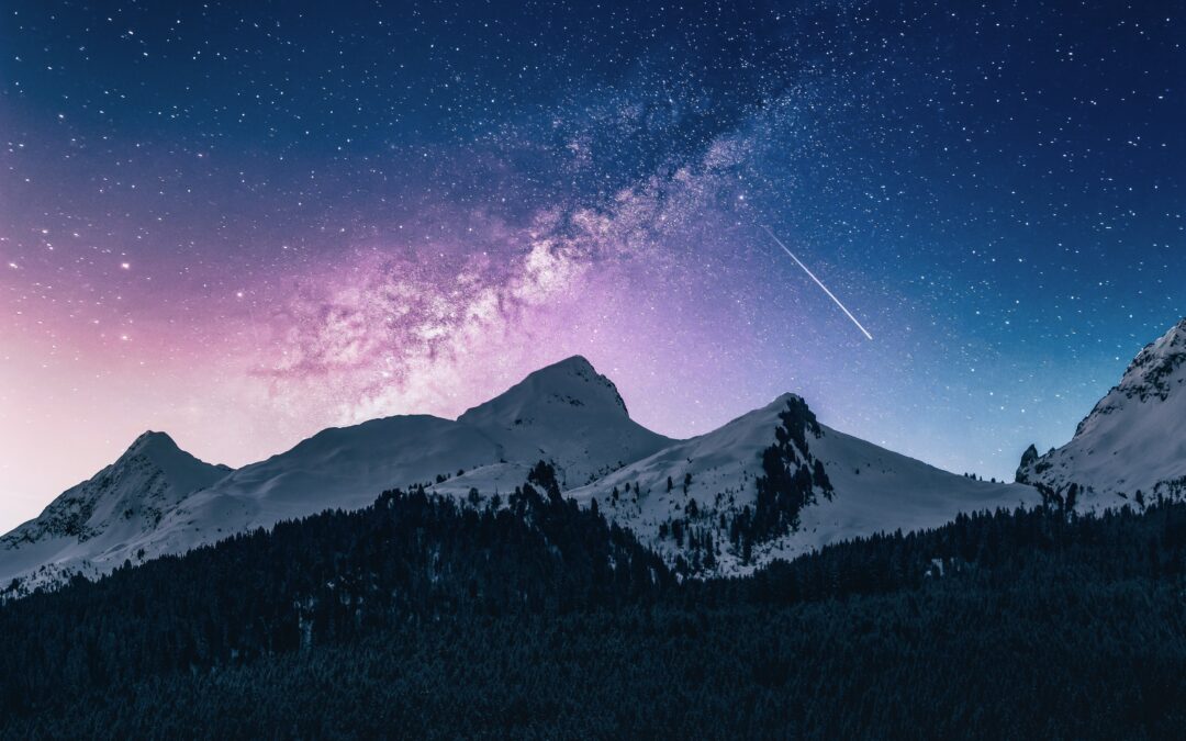 This is a photo by Benjamin Voros. It features a night sky set amongst a backdrop of mountains. There is a shooting star streaking through the night sky.